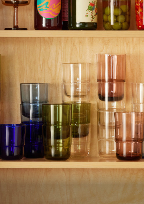 A wooden shelf displays colorful stacked glass tumblers in blue, green, clear, pink, and brown tones. The upper shelf holds jars, bottles, and a goblet.