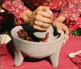 A person in a colorful, patterned shirt grinds dried red chilies in a gray stone molcajete on a red tablecloth. The persons hand is holding a pestle, crushing the chilies inside the bowl.