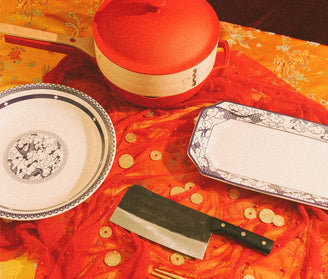 A traditional table setting with a red-lidded pot, two decorative plates, a large cleaver knife, and scattered gold coins on red and yellow patterned fabric.