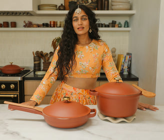 A woman in a yellow floral outfit stands in a modern kitchen, behind a counter with a terracotta-colored pan and pot. Shelves with dishes and spices are visible in the background.