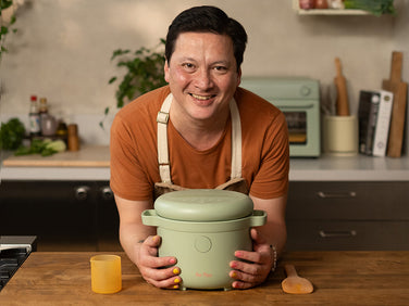 A person wearing an apron stands in a kitchen, smiling and holding a green cooking pot on a wooden counter with a cup and wooden spatula nearby.