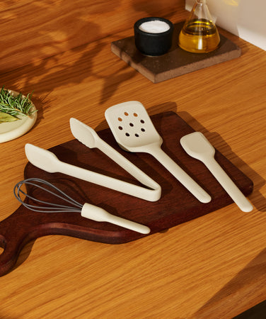 A set of light-colored silicone kitchen utensils, including a whisk, tongs, slotted turner, and spatula, arranged on a wooden cutting board.