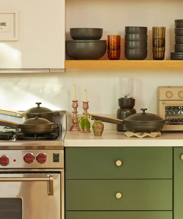 A modern kitchen with green cabinets, a stainless steel oven, cookware on the stove, and shelves displaying stacked dishes, glasses, and a small toaster. A framed fish drawing hangs on the wall. Sunlight brightens the scene.