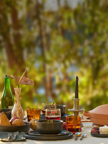 A beautifully set outdoor table with a terracotta pan in the center, surrounded by plates, glasses of drinks, fruits, candles, and vases with flowers, against a blurred natural background.