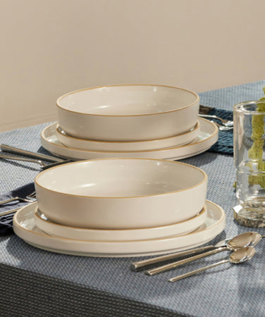 A neatly set dining table with white dishes, silver cutlery, clear glasses, and napkins on a blue tablecloth. Decorative items include a brown vase, a stone ring, and a green plant garland.