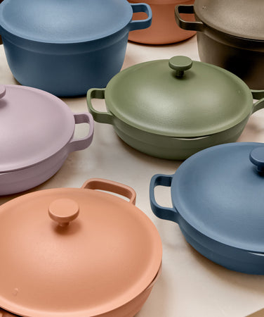 A collection of colorful Dutch ovens and pots with lids and handles, including green, blue, lavender, and peach tones, arranged on a countertop. A wooden spoon rests on one of the lids.