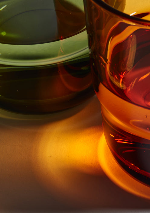 Close-up of two translucent glass cups, one green and one amber, with light passing through them, casting colorful reflections and shadows on a light surface.