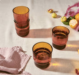 Four amber-tinted drinking glasses sit on a light tablecloth, two stacked and two separate. A textured pink cloth, citrus fruit, and purple shallots are scattered nearby in soft natural light.