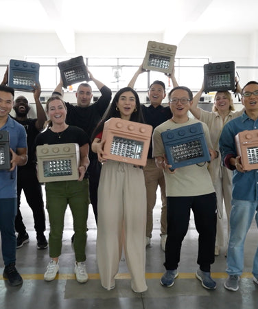 A group of people stand indoors smiling and holding colorful toaster ovens, with some raising the ovens above their heads. The setting appears to be a bright, spacious workshop or factory.