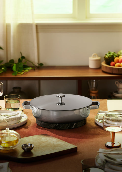 A covered stainless steel pot sits on a trivet at the center of a dining table set for a meal, with plates, glasses, and bowls arranged around it.