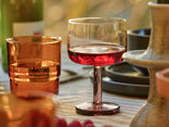 A close-up of a tabletop set for a meal, featuring a clear stemmed glass filled with red liquid and an amber-colored tumbler, with plates and a candle holder in the background.