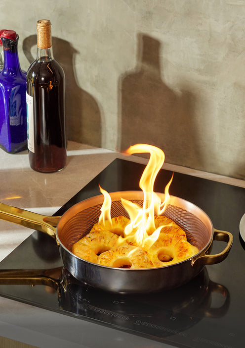 A frying pan on a stovetop contains pineapple slices that are being flambéed, with bright flames rising from the fruit. A wine bottle and a blue bottle are in the background on the counter.
