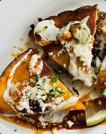 Two slices of cheesy, saucy birria quesadillas topped with crumbled cheese, chopped onions, cilantro, and crema on a white plate, with a lime wedge on the side.