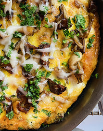 A close-up of a mushroom and herb frittata in a skillet, topped with melted cheese, sautéed mushrooms, and chopped fresh parsley.
