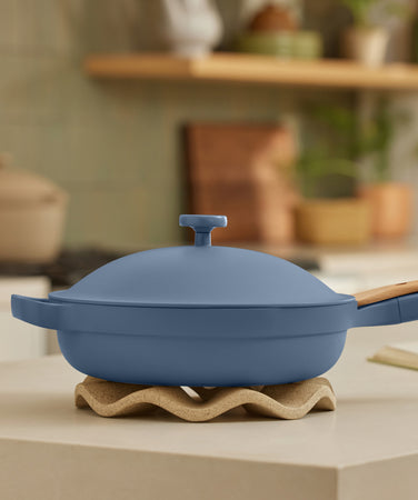 A blue nonstick pan with a lid and wooden handle sits on a wavy trivet on a kitchen counter; the background shows a blurred kitchen with a pot, plant, and an open book.