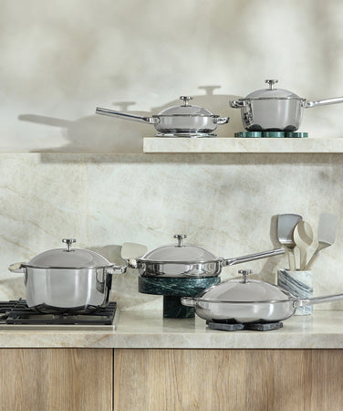 A set of stainless steel pots and pans with lids sits on a modern gas stove, with utensils on the marble countertop nearby. The background features a light-colored stone backsplash and wooden cabinetry.
