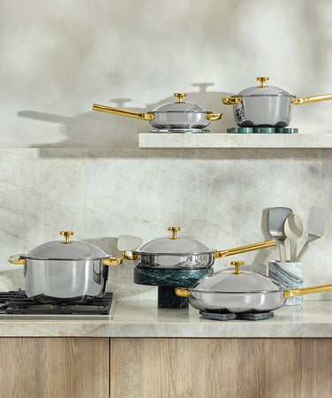 Two stainless steel pots with gold handles and lids sit on a gas stove. In the background, utensils are in a marble holder next to a wooden cutting board. The scene is a modern kitchen countertop.
