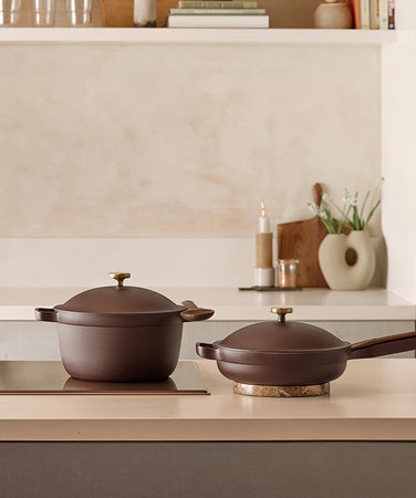 Two brown cookware pieces, a pot with a lid and handles, and a skillet with a long handle and lid, sit on a light kitchen counter with minimalist decor in the background.