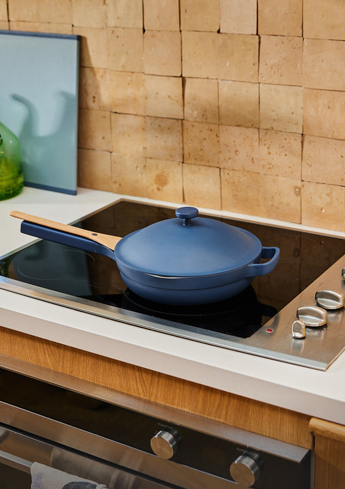A blue lidded skillet with a wooden handle sits on a black electric stovetop in a modern kitchen with a tan tiled backsplash and an oven below.