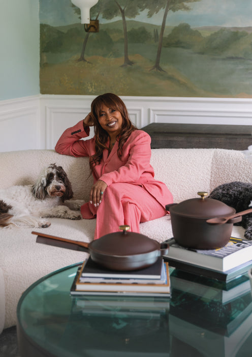A woman in a pink suit sits on a cream sofa, smiling with her hand on her head. Two dogs rest beside her. A glass table with cookware and books is in the foreground. A mural of trees decorates the wall behind her.