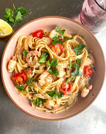 A bowl of creamy pasta with shrimp, cherry tomatoes, and fresh parsley, garnished with black pepper. A lemon wedge, parsley sprig, and a glass of water are beside the bowl on a metallic surface.