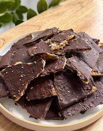 A plate filled with irregular pieces of chocolate-covered toffee topped with sea salt, sitting on a wooden table with green leaves blurred in the background.