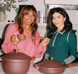 Two women stand in a kitchen smiling at the camera. One woman in a pink outfit holds a large brown spoon. In front of them are two brown pots with gold handles on a white counter. Shelves with books and plants are behind them.