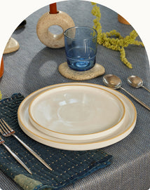 A set table with two stacked cream plates, a blue glass, two spoons, a fork, and a knife on a navy textured napkin over a blue-gray tablecloth. Decorative ceramic objects and yellow-green flowers are in the background.