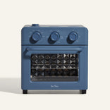 A compact blue countertop oven with three control dials on top, a pull-down glass door, and a geometric pattern on the inside of the door. The brand name Our Place is visible below the window.