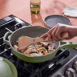 A person stirs a variety of mushrooms in a green skillet on a stovetop using a wooden spatula. A glass and plate are on the counter nearby.