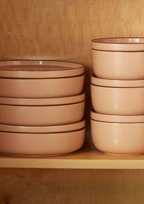 A stack of pink ceramic plates sits next to a stack of matching pink bowls on a wooden shelf against a wooden background.