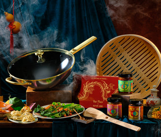 A steaming wok with a gold handle, a bamboo steamer, jars of chili sauces, dumplings, leafy greens, and a red box with a dragon, set against a dark, dramatic fabric backdrop with traditional Asian decor.