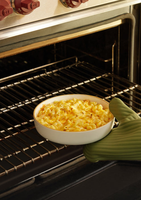 A hand wearing a green oven mitt is placing a white dish of baked macaroni and cheese into an open oven with red knobs.