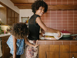 A smiling woman lifts a pot lid on the stove in a cozy kitchen, while a child with similar curly hair leans forward beside her, both facing away from the camera.