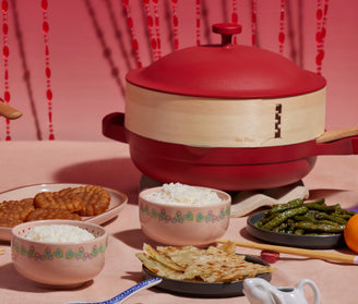 A red pot with wooden handles sits on a table set with bowls of white rice, crispy green beans, scallion pancakes, and pieces of fried food, against a pink backdrop with red bead strings.