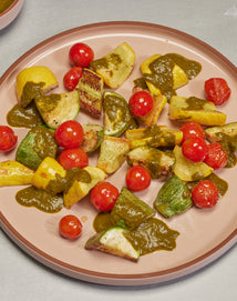A plate of roasted vegetables, including yellow squash, zucchini, and cherry tomatoes, topped with a green sauce, arranged on a light pink plate.