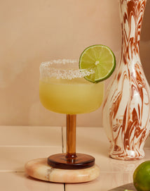 A margarita with a salted rim and a lime wheel garnish sits on a marble coaster. The drink is served in a brown-stemmed glass, with a marbled vase and part of a lime in the background.