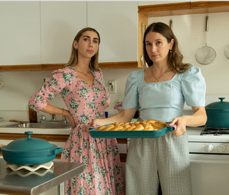 Two women in vintage-style dresses stand in a kitchen. One holds a baking tray of golden bread rolls, while the other stands beside her. The kitchen has white cabinets and teal cookware.