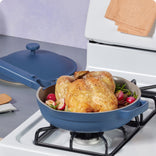 A whole roasted chicken with radishes and herbs sits in a blue pan on a stovetop, with the matching blue lid resting nearby. A beige towel is on the stove in the background.