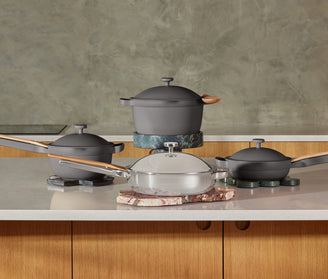 A set of modern cookware including two gray pots, a gray pan, and a silver pan with wooden handles, arranged on a kitchen counter with a greenish textured wall in the background.