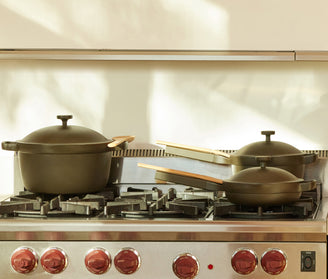 A set of matte black pots and pans with wooden handles sits on a stainless steel gas stove with red knobs in a sunlit kitchen.