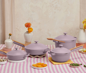 Lavender-colored cookware set with wooden handles displayed on a pink and white striped tablecloth, surrounded by cut citrus, small vases with flowers, and a beige background.