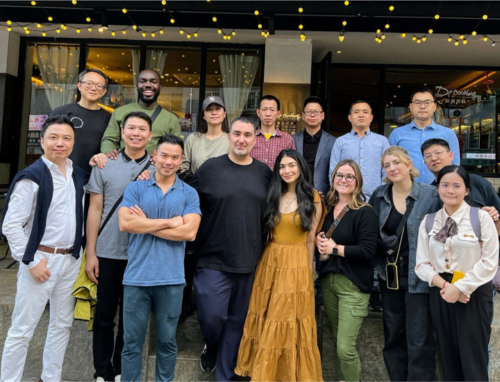 A group of sixteen people pose together and smile outside a restaurant. Some are standing, others are kneeling in front. Warm yellow string lights hang overhead, and the restaurant entrance is visible in the background.