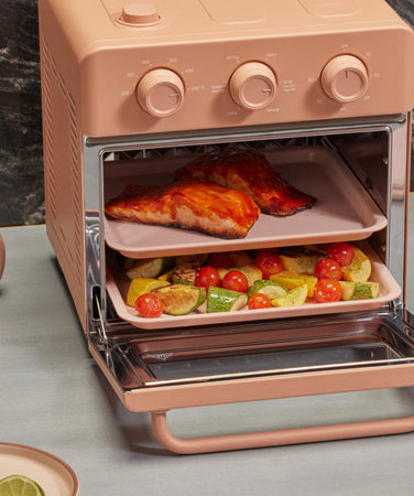 A pink countertop oven with its door open, showing salmon fillets on the top rack and chopped vegetables with cherry tomatoes on the bottom rack. A bowl, onion, and lime wedges are nearby on a gray surface.