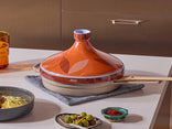 A ceramic tagine with a patterned orange lid sits on a cloth trivet on a kitchen counter, surrounded by small dishes of olives, spices, and a bowl of food.