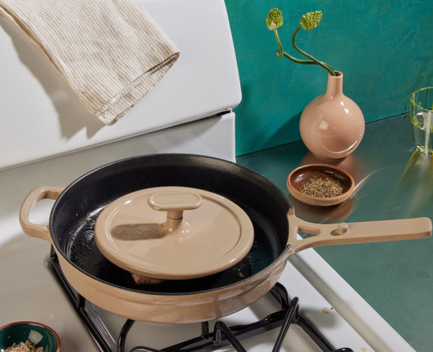 A beige skillet with a matching lid sits on a stove. Nearby are a pink vase with green stems, a glass of water, a striped towel, and bowls with spices on a green countertop.