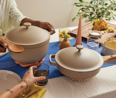 Two people sit at a table set with beige cookware, a blue table runner, a lit candle, glasses of water, and a cutting board with roasted cauliflower and sauce. One person holds a pot, another reaches for a glass.