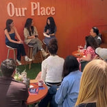 A small group watches a panel of three women in conversation on stools in front of an orange wall with Our Place written on it. Drinks and notebooks are on the tables.