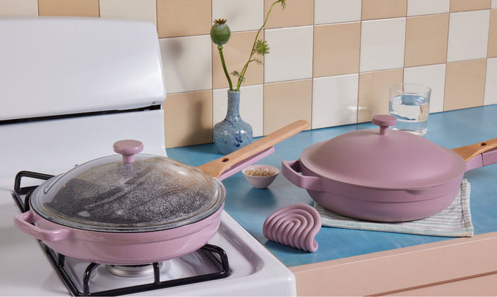 Translation missing: en.Two pink pans with wooden handles are on a kitchen stove and counter; one is covered with a glass lid steaming, the other is closed. A blue vase with flowers, a glass of water, pot holder, and bowl are in the background