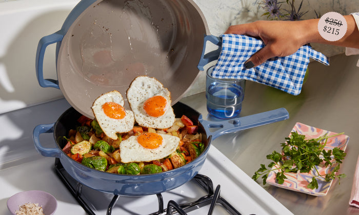 Translation missing: en.A hand holding a blue skillet with a checked towel slides three sunny-side-up eggs onto a pan of sautéed vegetables on a stove. Price tag in corner shows a discount from $250 to $215. Fresh herbs and a glass of water nearby